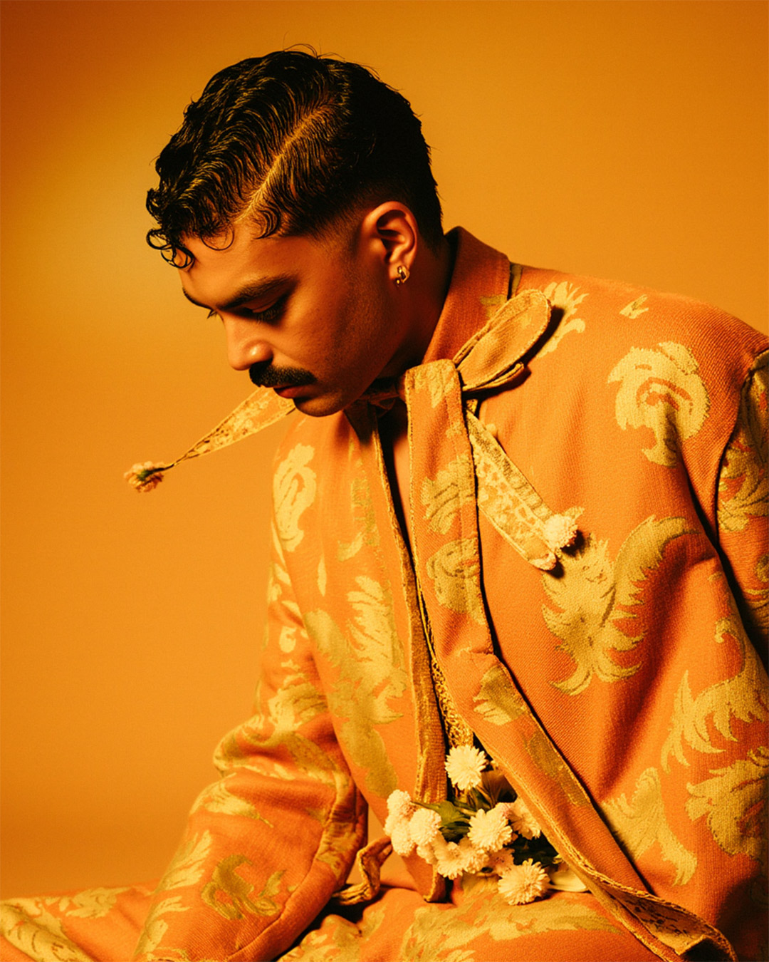 Marco Vincit veste um traje brocado laranja com detalhes dourados. A iluminação quente realça sua expressão introspectiva, criando um visual sofisticado e atemporal. — Laranja, Marco Vincit