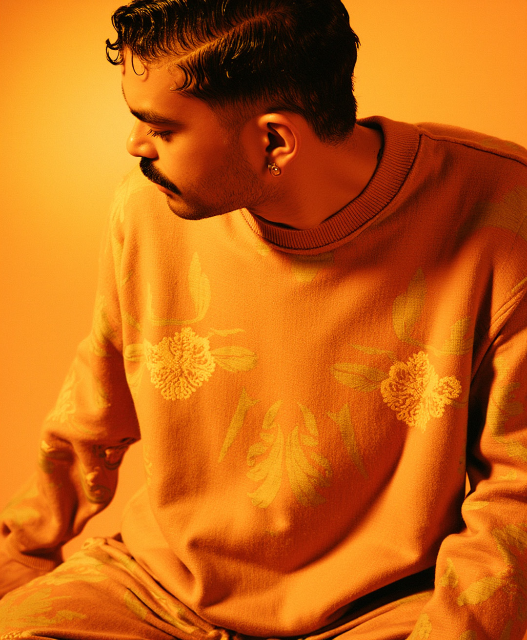 O artista usa um moletom laranja com bordados florais dourados, destacando a fusão entre moda e arte com uma atmosfera aconchegante. — Laranja, Marco Vincit