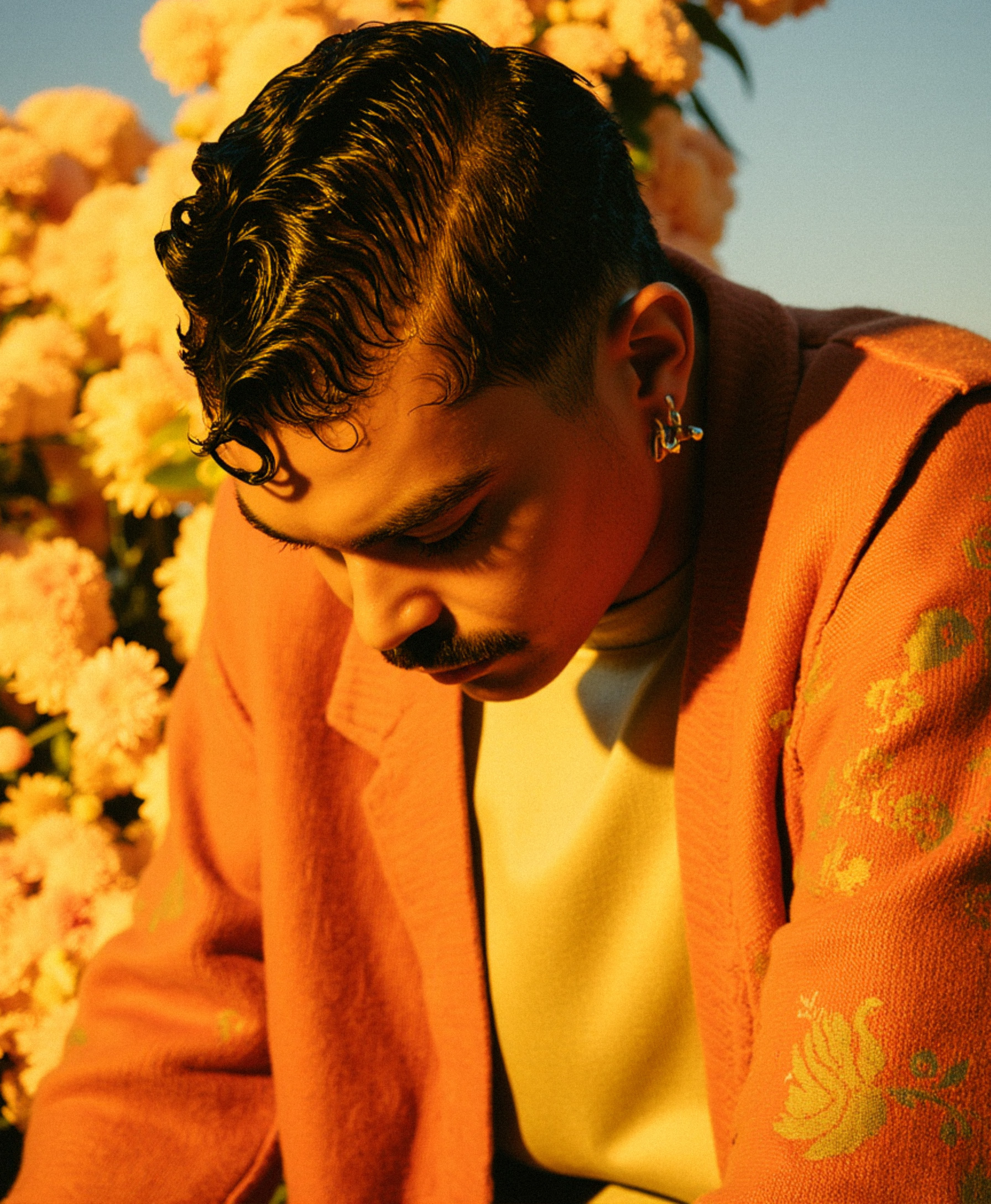 Cercado por flores em tons pastel, Marco Vincit usa um casaco laranja com estampas florais. A paleta quente reforça a estética romântica e nostálgica do ensaio. — Laranja, Marco Vincit