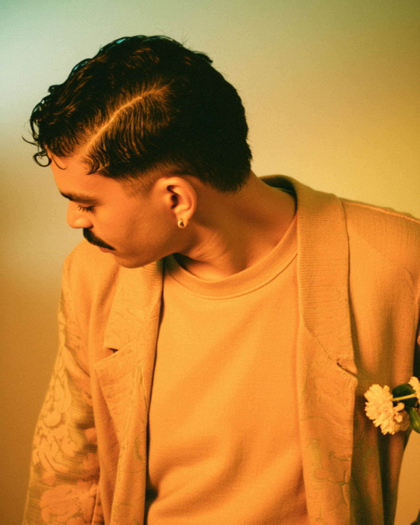 O artista veste um suéter laranja decorado com flores brancas. A composição simples, porém impactante, destaca sua identidade visual única. — Laranja, Marco Vincit
