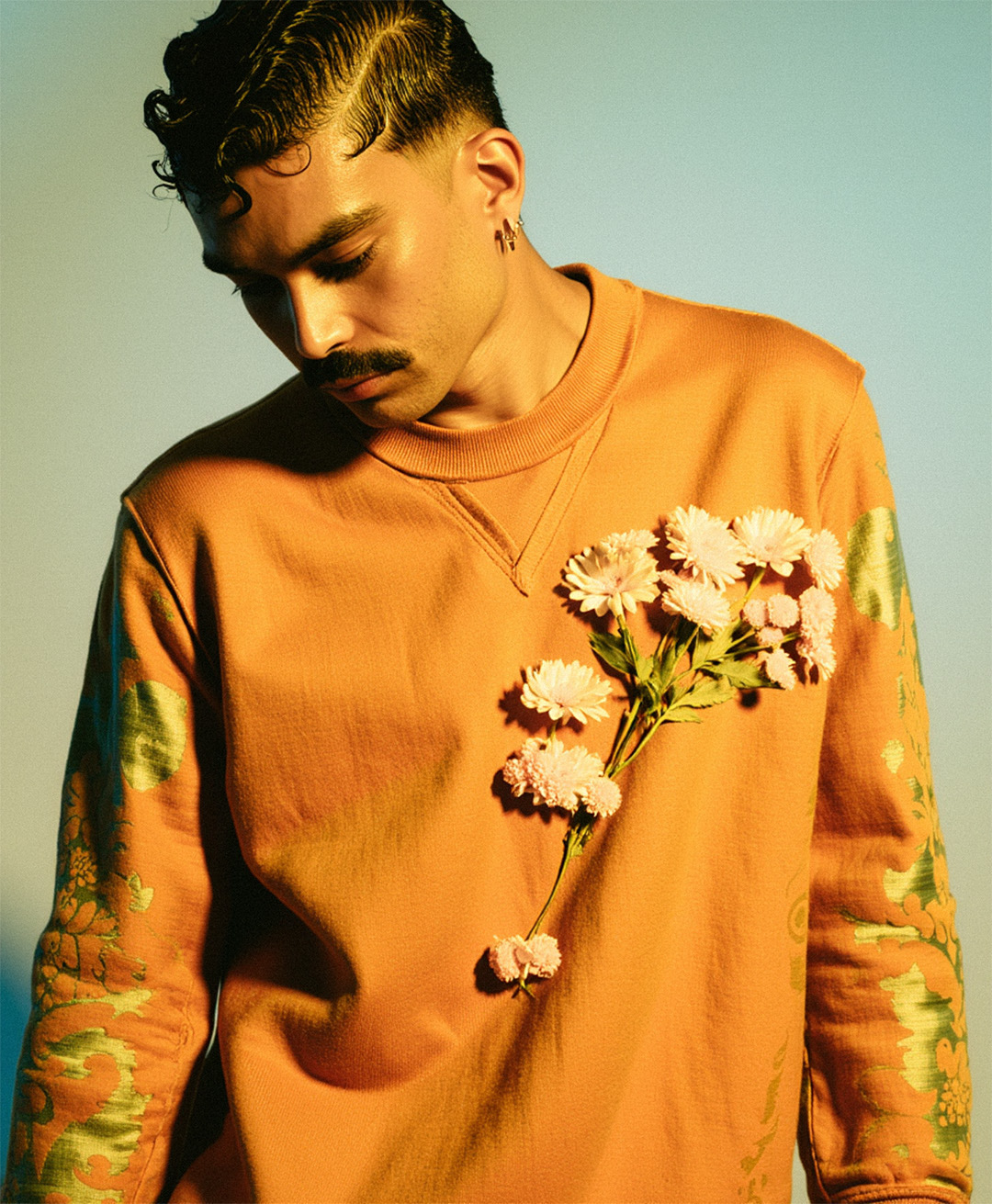 Marco Vincit segura um arranjo floral branco contra seu moletom laranja. O contraste entre os elementos naturais e a cor intensa cria um visual marcante. — Laranja, Marco Vincit