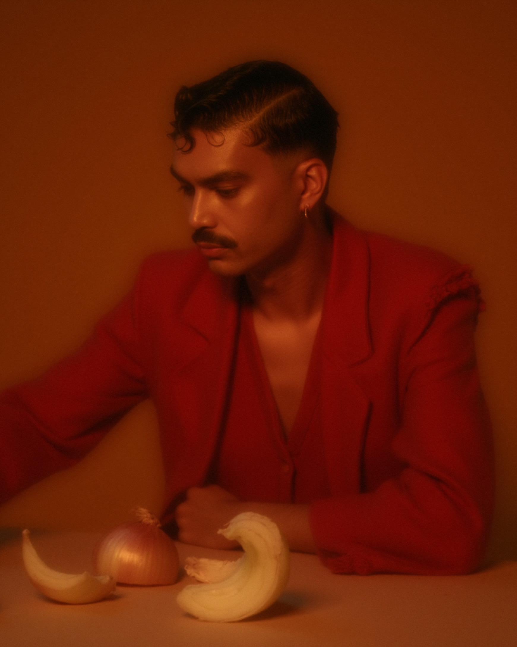 Marco Vincit posa com um blazer vermelho vibrante em um cenário suavemente iluminado. Elementos simbólicos como cebolas descascadas adicionam uma camada de narrativa e mistério à composição. — Retratos, Marco Vincit