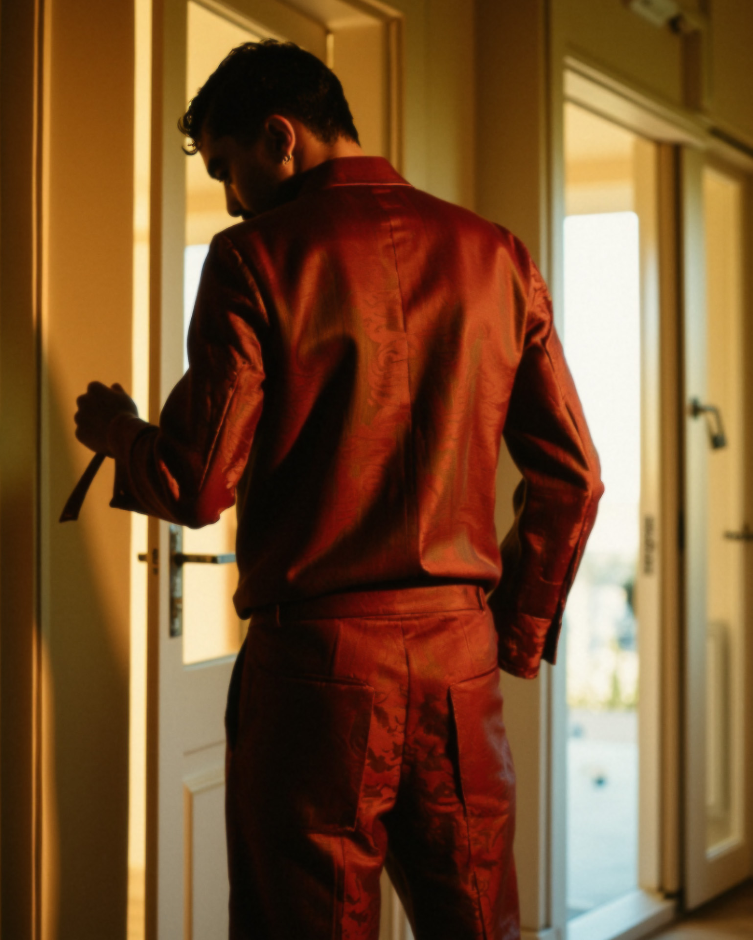 Marco Vincit de costas, vestindo um traje vermelho sofisticado enquanto segura uma alça de bolsa. A iluminação dourada e a composição sugerem um momento de transição e introspecção. — Retratos, Marco Vincit