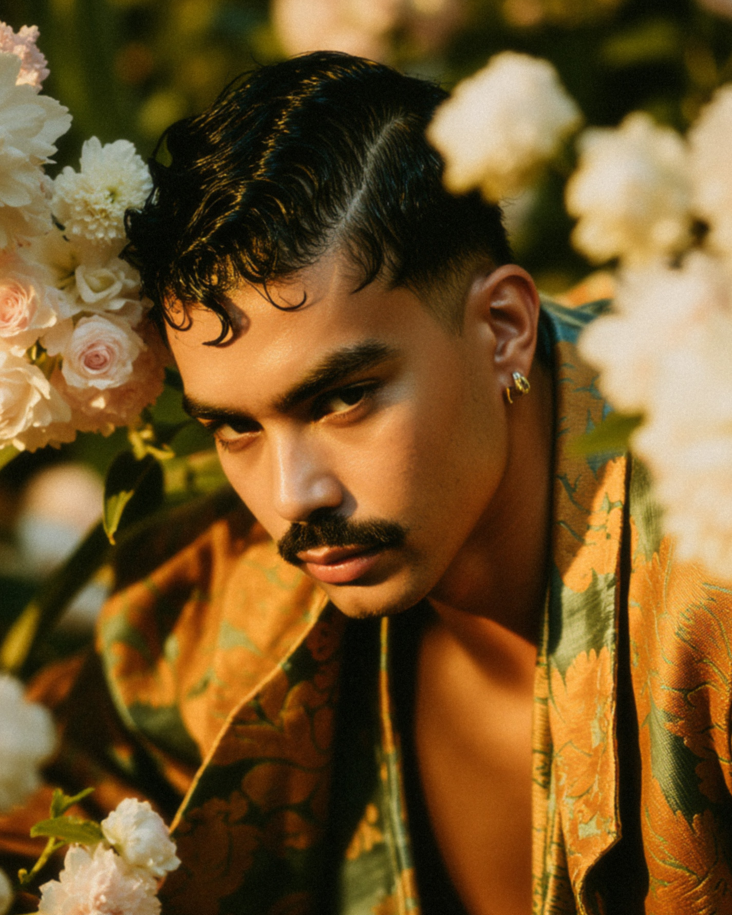 Marco Vincit posa entre flores brancas e rosas, vestindo um terno com padrões dourados e verdes. A luz suave realça sua expressão intensa, criando um contraste entre delicadeza e força. — Retratos, Marco Vincit