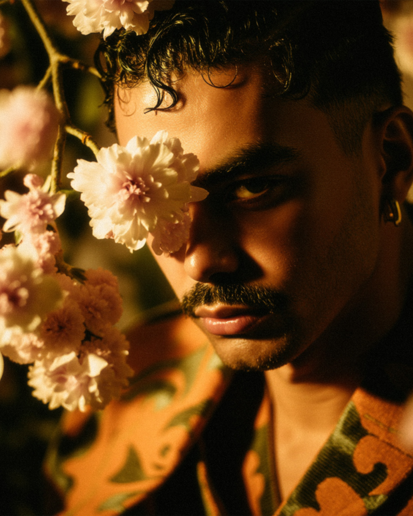 Marco Vincit surge parcialmente oculto por flores brancas, com um olhar intenso e penetrante. A iluminação dourada e as sombras dramáticas adicionam profundidade à composição, criando um efeito artístico e enigmático. — Retratos, Marco Vincit