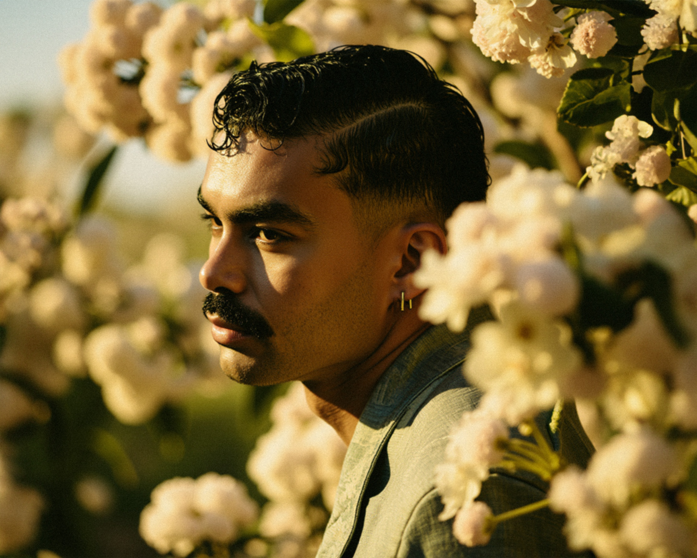 Marco Vincit posa cercado por flores brancas sob a luz quente do entardecer. Seu olhar contemplativo e a paleta de tons suaves criam uma atmosfera serena e poética. — Retratos, Marco Vincit
