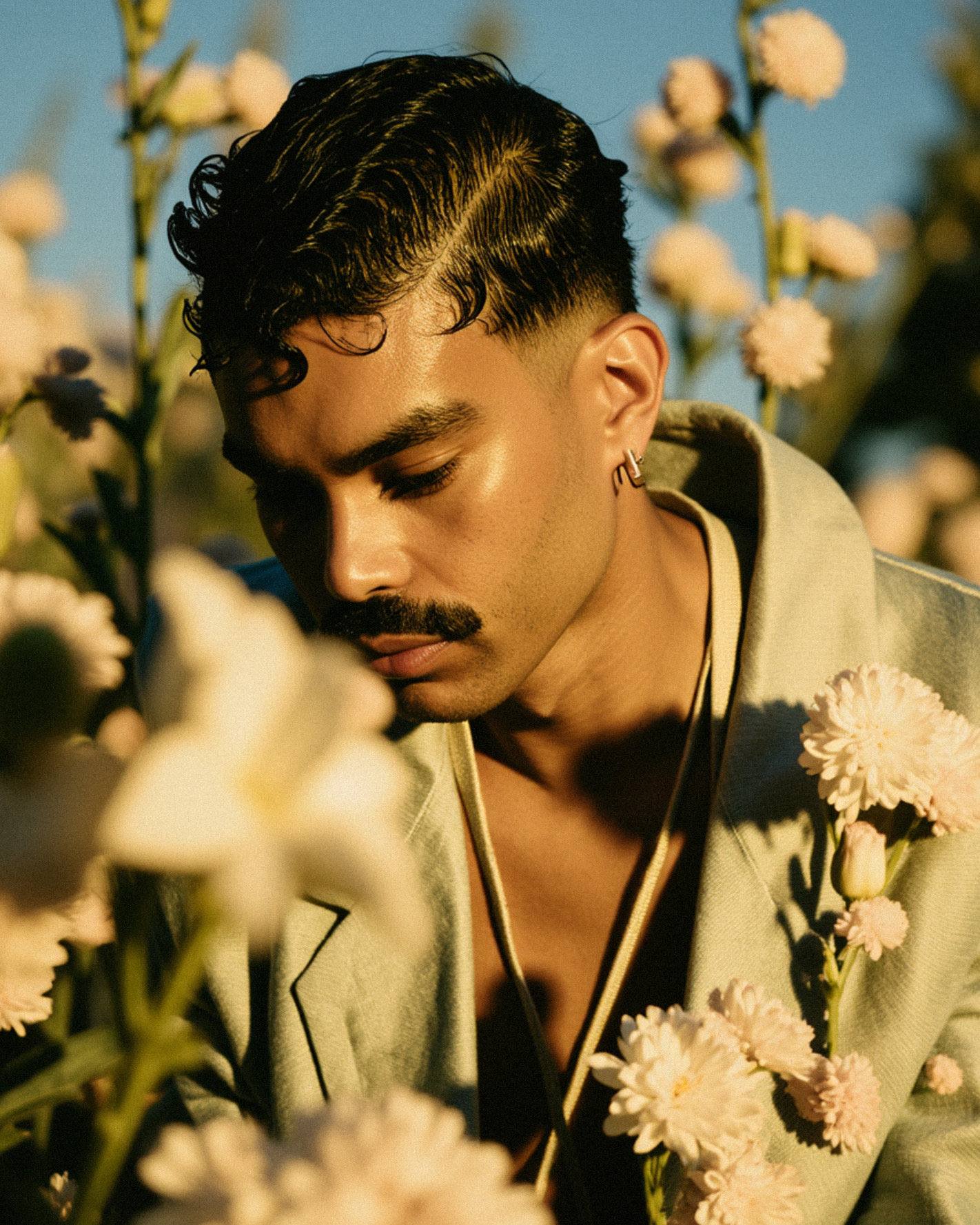 Marco Vincit veste um blazer verde claro adornado com flores, cercado por um campo dourado ao entardecer. A luz suave e seu olhar introspectivo criam uma atmosfera de tranquilidade e conexão com a natureza. — Retratos, Marco Vincit