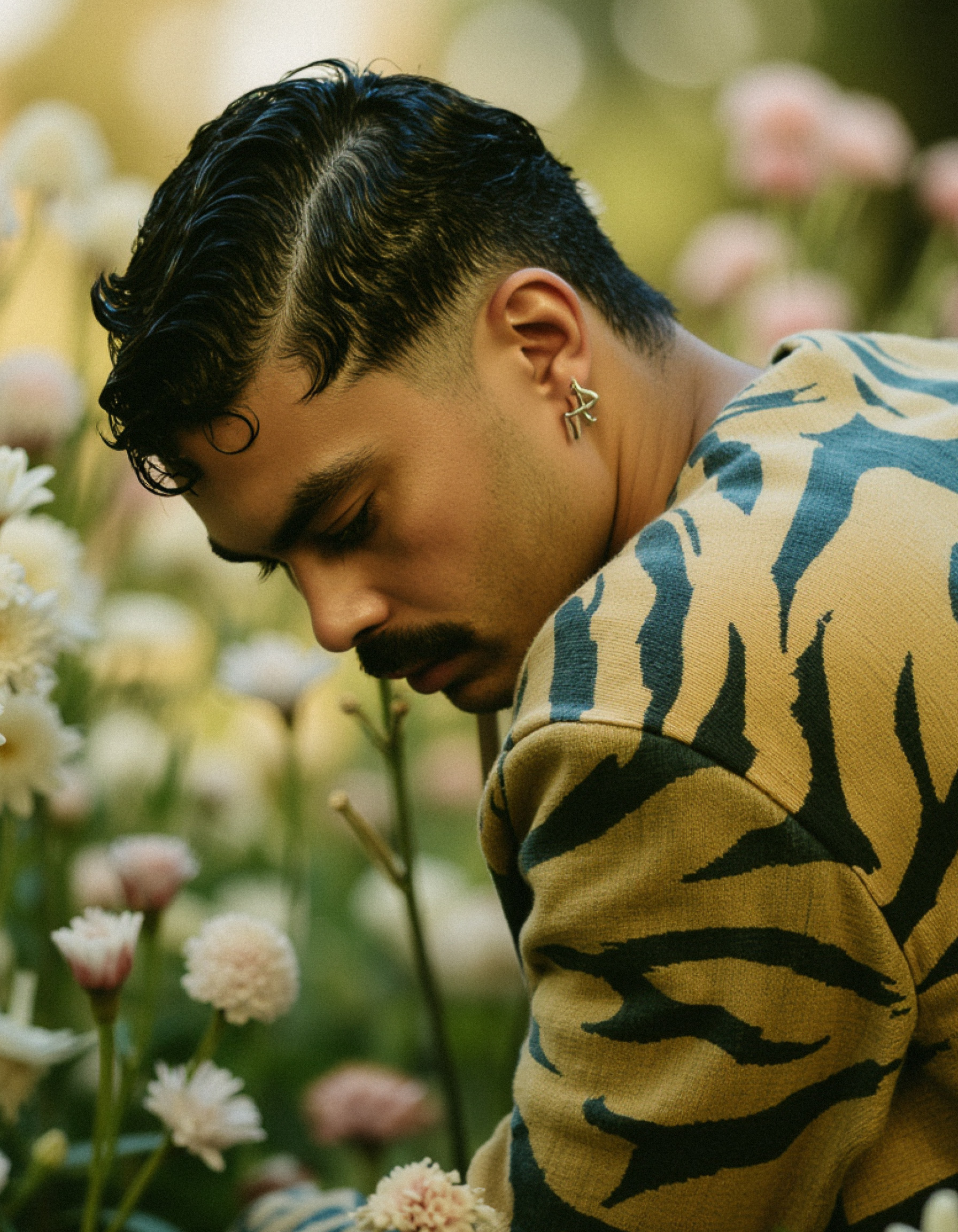 Marco Vincit veste um suéter com padrão orgânico enquanto interage com flores brancas e rosas. Seu olhar introspectivo e a luz suave criam uma cena serena e harmoniosa. — Retratos, Marco Vincit