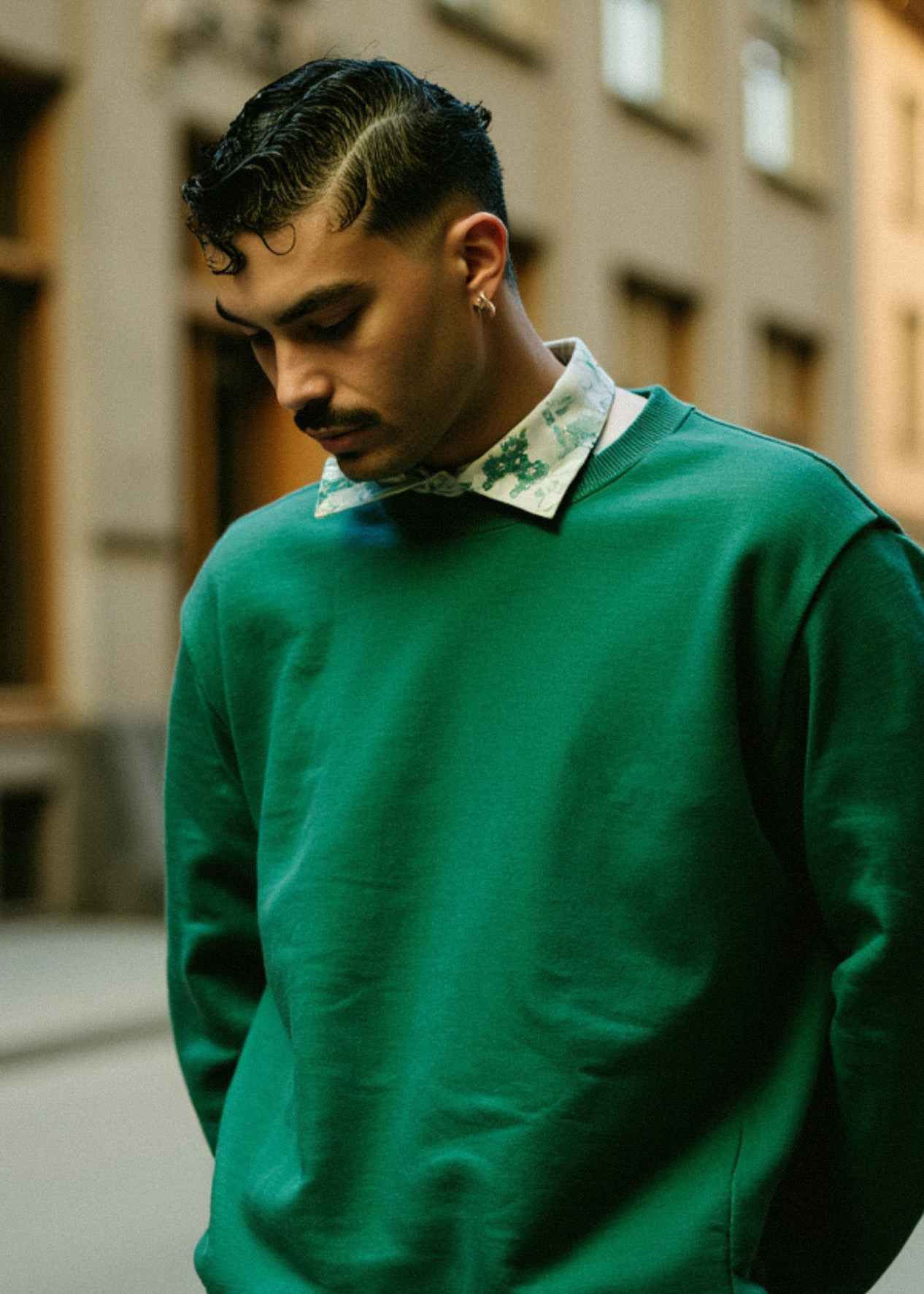 Marco Vincit veste um suéter verde vibrante com camisa estampada, posando em um cenário urbano. Seu olhar introspectivo e a iluminação suave criam uma atmosfera moderna e sofisticada. — Retratos, Marco Vincit