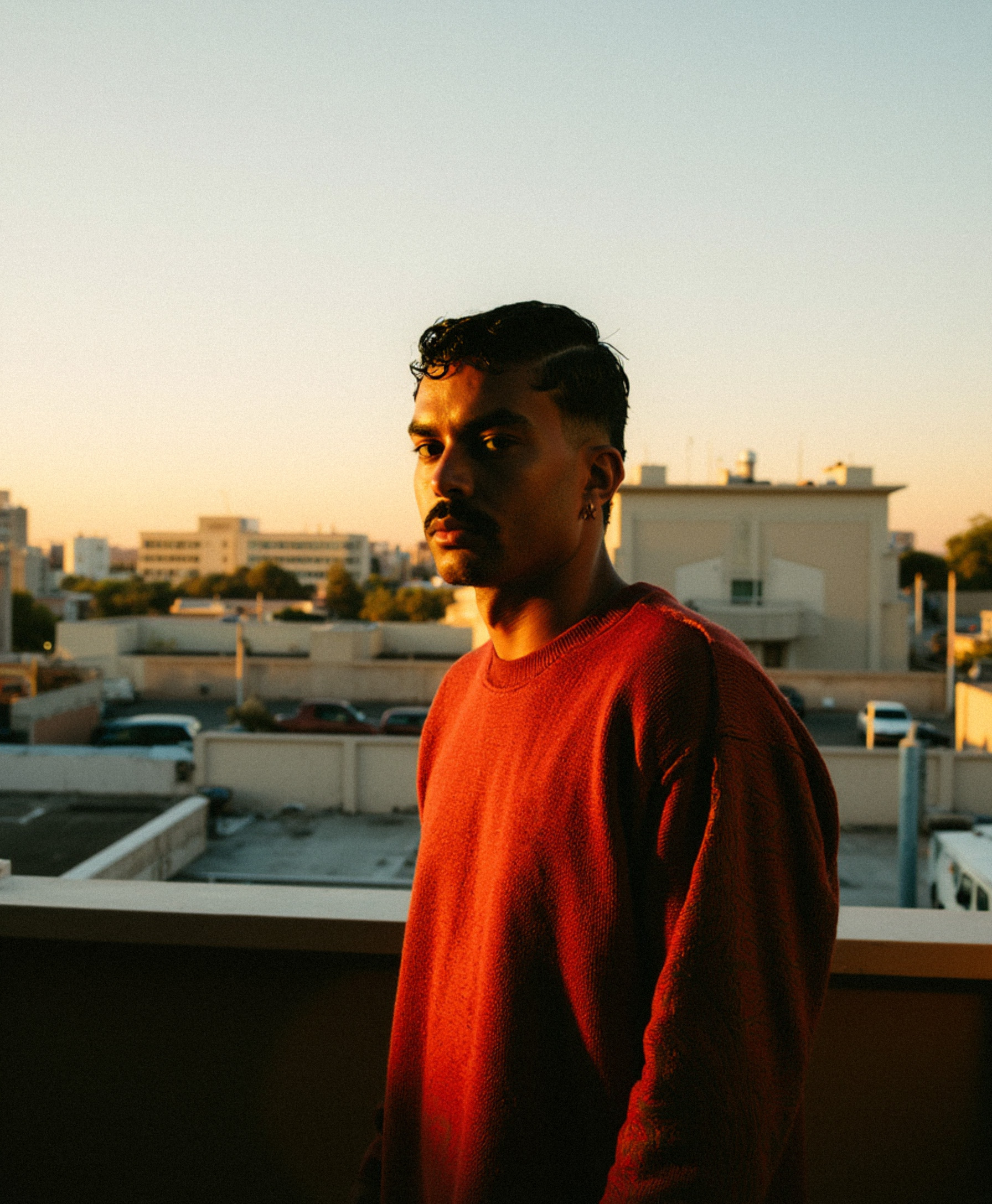 Marco Vincit veste um suéter vermelho enquanto a luz dourada do pôr do sol ilumina seu rosto. A cidade ao fundo adiciona um toque melancólico e cinematográfico à composição. — Retratos, Marco Vincit