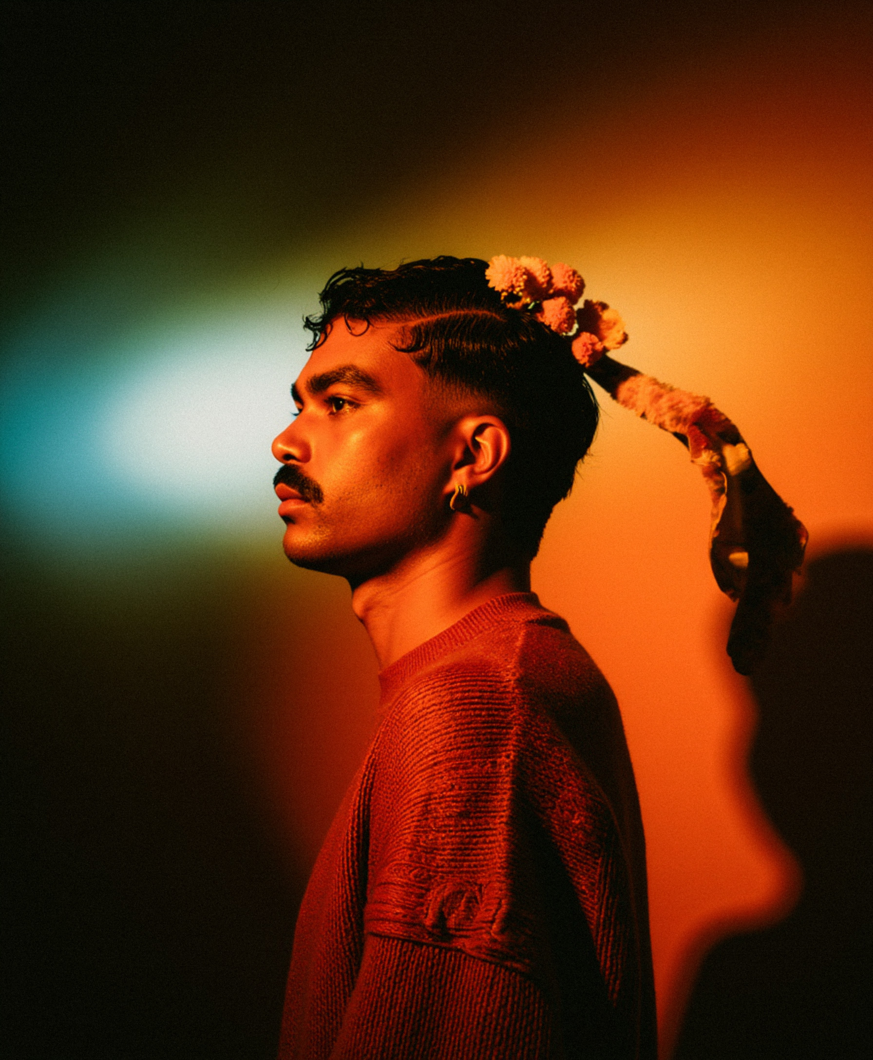Marco Vincit posa de perfil com um suéter vermelho e um acessório floral no cabelo. A iluminação multicolorida cria um efeito dramático e surreal na composição. — Retratos, Marco Vincit