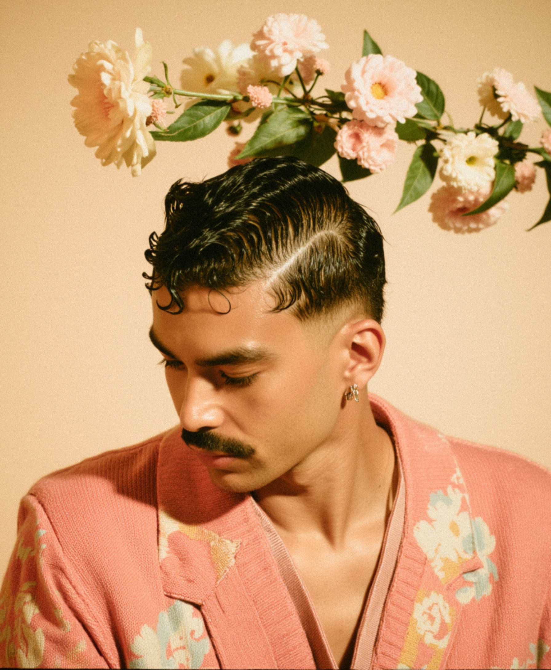 Marco Vincit veste um blazer rosa com detalhes florais, enquanto flores delicadas se entrelaçam ao fundo. A iluminação suave e a paleta pastel criam uma estética etérea e romântica. — Retratos, Marco Vincit