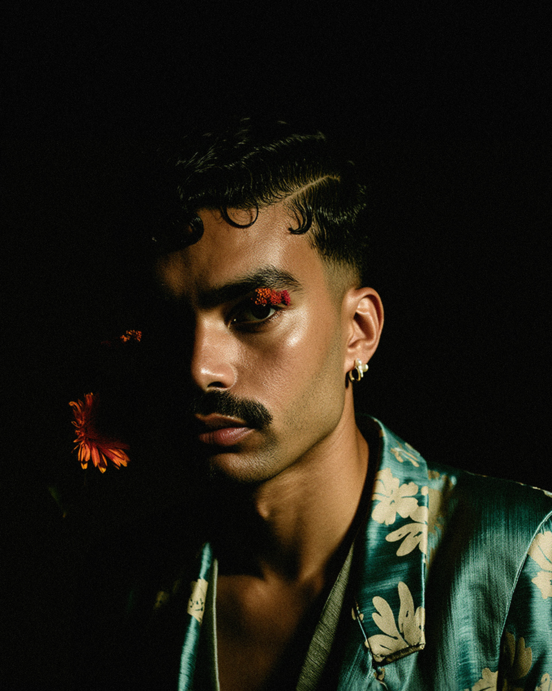 Marco Vincit veste um blazer de cetim azul com estampas florais, enquanto a iluminação dramática destaca metade de seu rosto. Pequenas flores vermelhas adornam seu olhar, criando um contraste intenso e enigmático. — Retratos, Marco Vincit