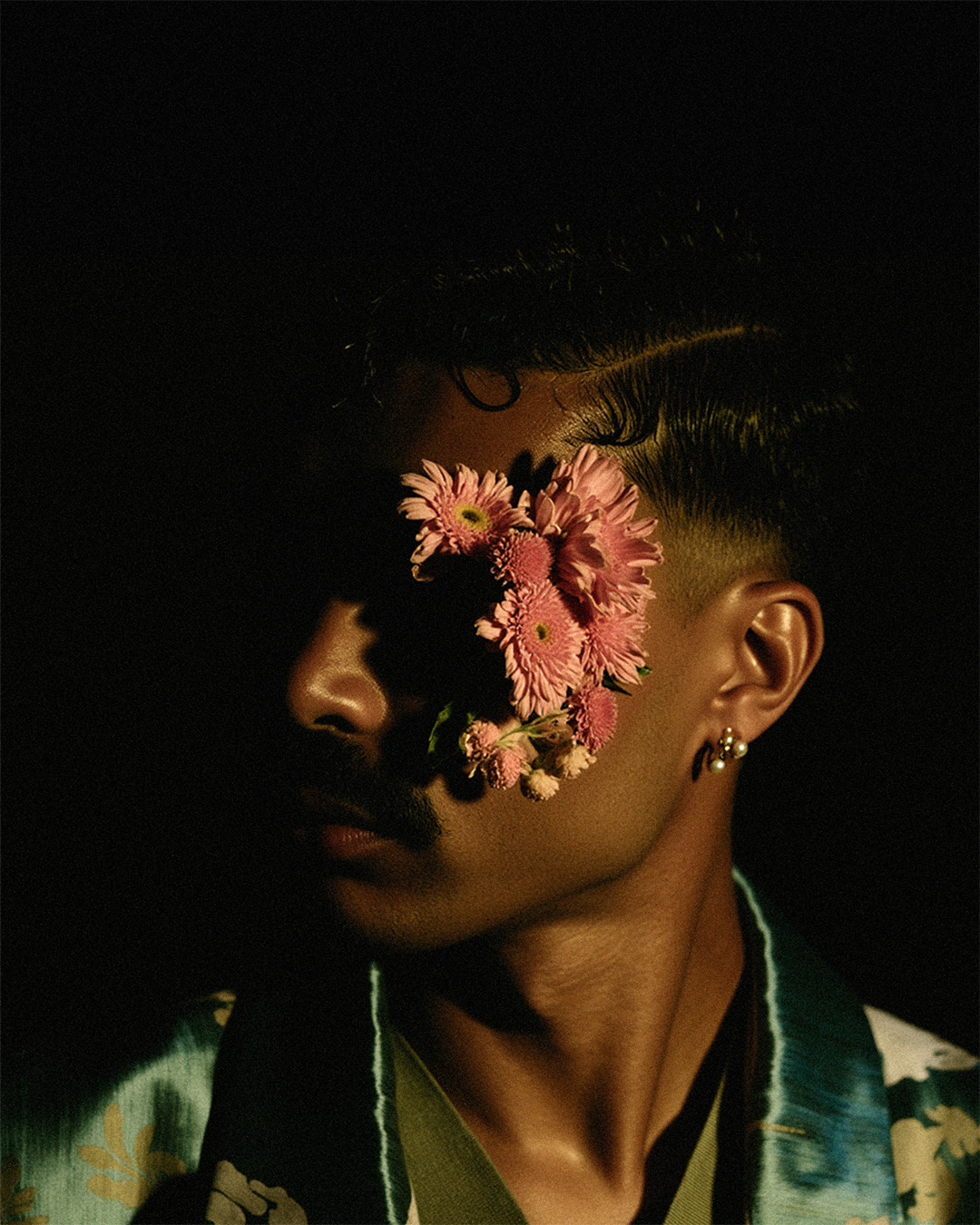 Marco Vincit surge na penumbra com flores cobrindo parte de seu rosto, criando um contraste entre luz e sombra. A iluminação sutil reforça a atmosfera misteriosa e poética da imagem. — Retratos, Marco Vincit