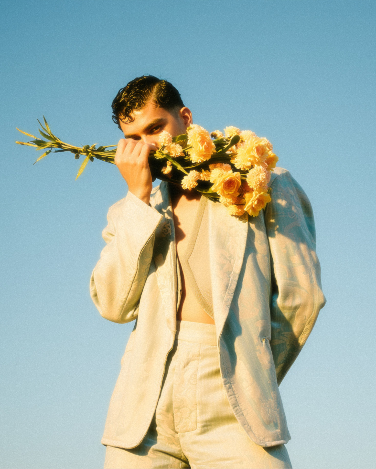 Marco Vincit veste um terno azul claro enquanto segura um buquê de flores amarelas contra o rosto. A luz suave e o fundo azul intenso criam um visual etéreo e elegante. — Rosas Amarelas, Marco Vincit