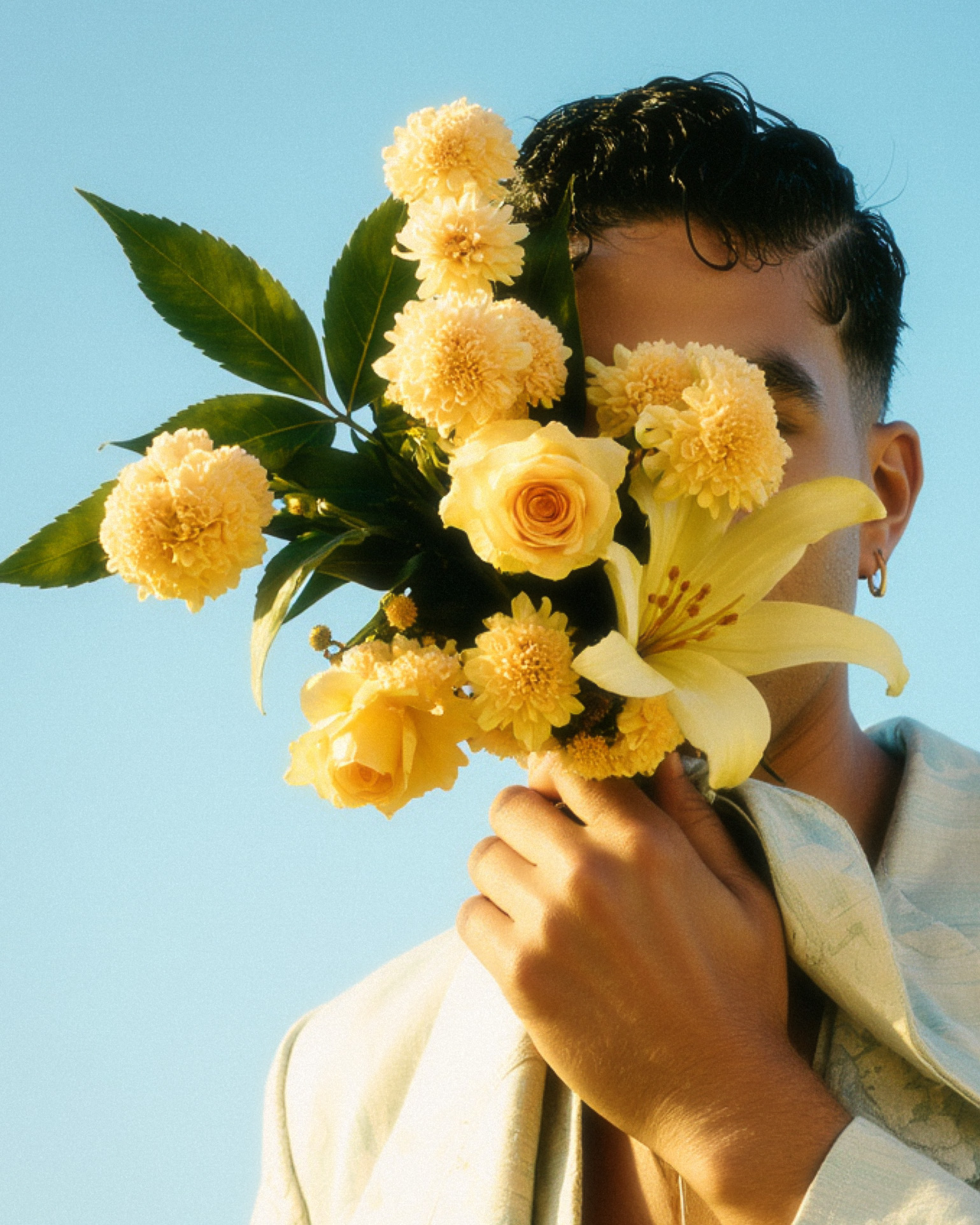 Marco Vincit segura um buquê de flores amarelas, cobrindo parcialmente seu rosto. O fundo azul vibrante e a iluminação suave criam um contraste elegante e poético. — Rosas Amarelas, Marco Vincit